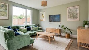A living room with a green sofa, a wooden coffee table, and a small cabinet. There are framed pictures on the wall and a window with a view of greenery outside.