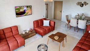 A modern living room with a red sofa set, a wooden coffee table, and a dining area with a white table and chairs. - 5mn from the beach, elegant relaxation apartment, Swimming pool, Playground, Park (Âïn-Harrouda)