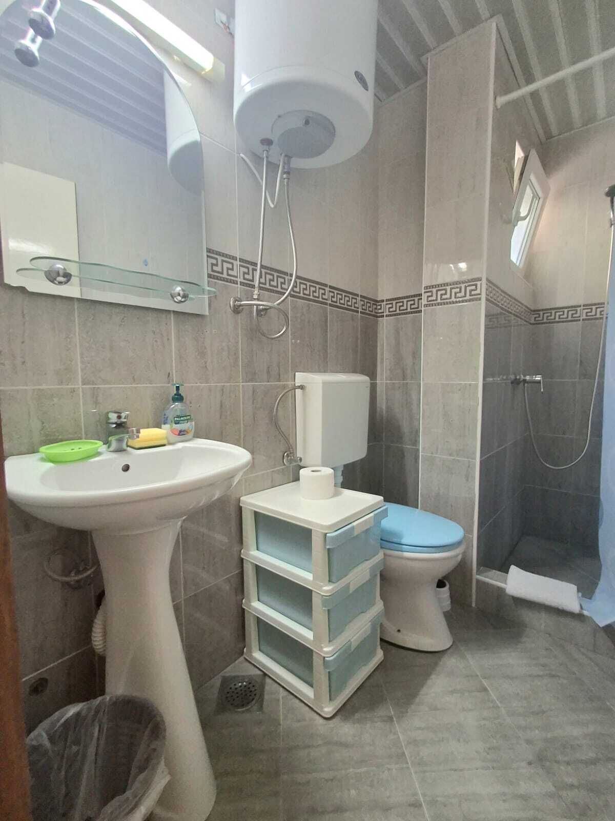 A bathroom with a pedestal sink, toilet, and shower. There are storage drawers under the sink and a trash bin.