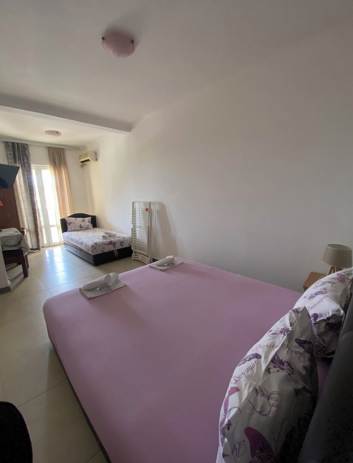 A hotel room with a large bed, a smaller bed, a desk, a chair, a lamp, and a window with curtains.