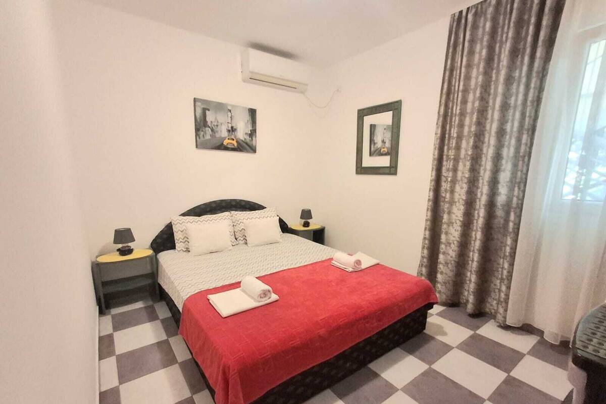 Ein Hotelzimmer mit einem Schachbrettmuster auf dem Boden, einem Bett mit roter Decke, zwei Nachttischen mit Lampen und zwei gerahmten Bildern an den Wänden.