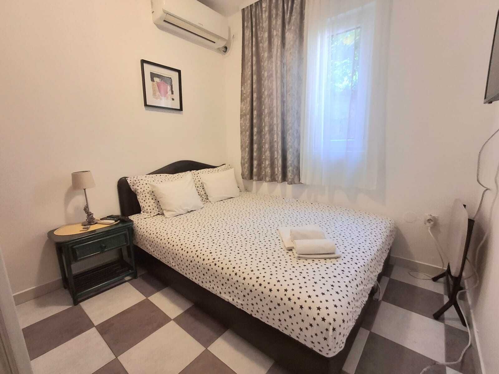 Quarto com cama, mesa de cabeceira, abajur, janela com cortinas e ar-condicionado fixado na parede.