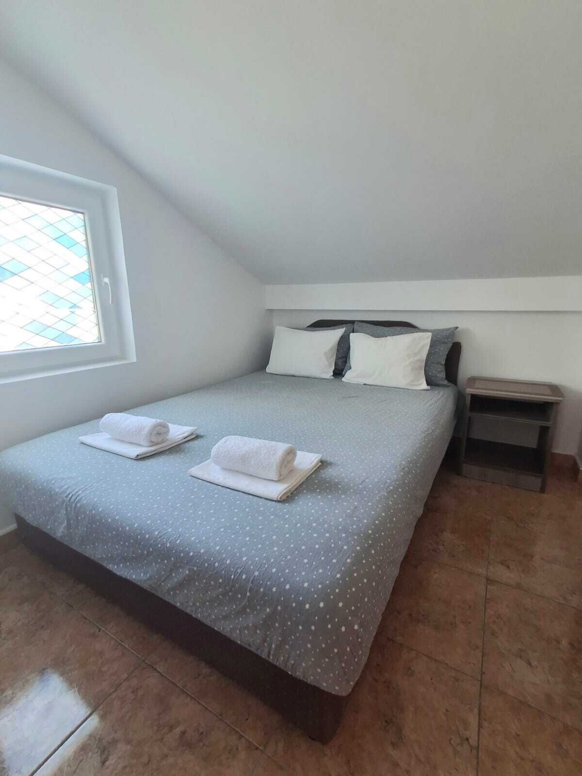 A neatly made bed with two rolled towels, a small bedside table, and a window with a view of a blue-tiled area.