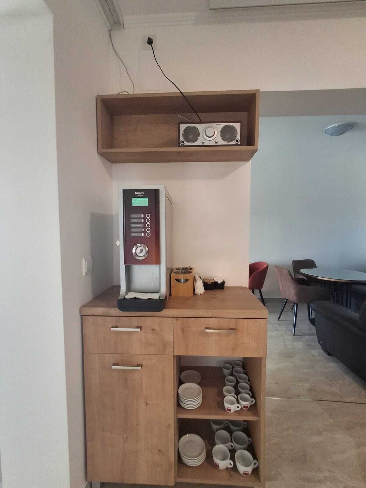 A kitchenette with a coffee machine, a shelf with speakers, and a dining area with a table and chairs.