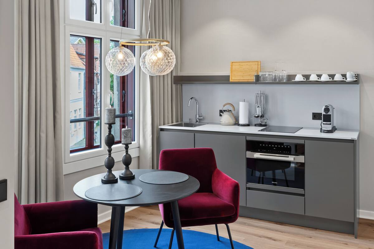 A modern kitchen with a dining area, including a round table, red chairs, and pendant lights.