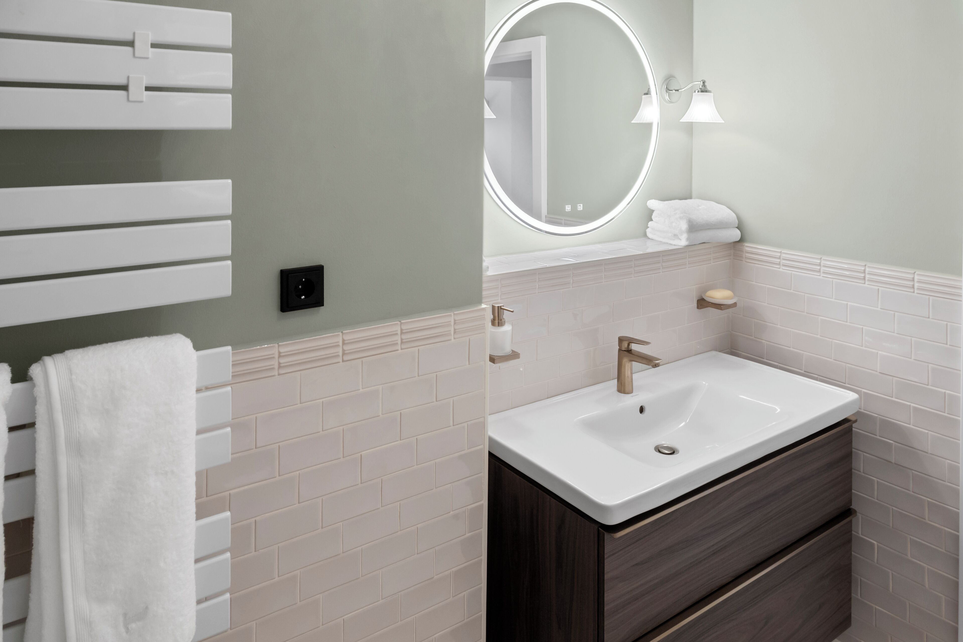Un baño con lavamanos blanco, tocador de madera oscura, toallero blanco y un espejo redondo con luces.