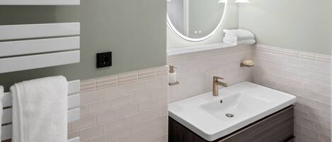Un baño con lavamanos blanco, tocador de madera oscura, toallero blanco y un espejo redondo con luces.