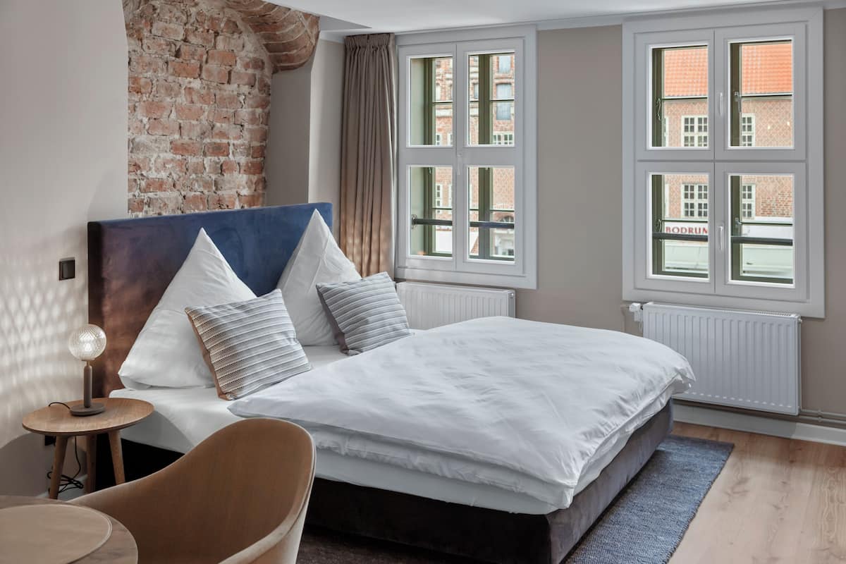 A bedroom with a large bed, a headboard, a nightstand, a chair, and a window with a view of buildings.