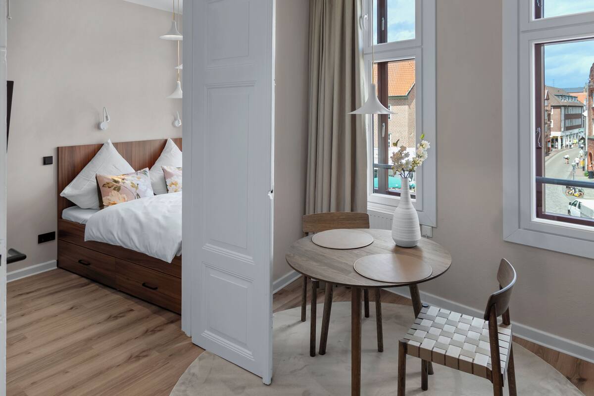 A bedroom with a bed, bedside tables, a round dining table with chairs, and a window with a view of buildings.