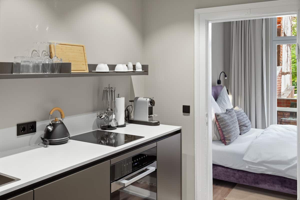 A modern kitchen with a built-in oven, a kettle, and a coffee maker. There is a bed with a purple headboard in the adjacent room.