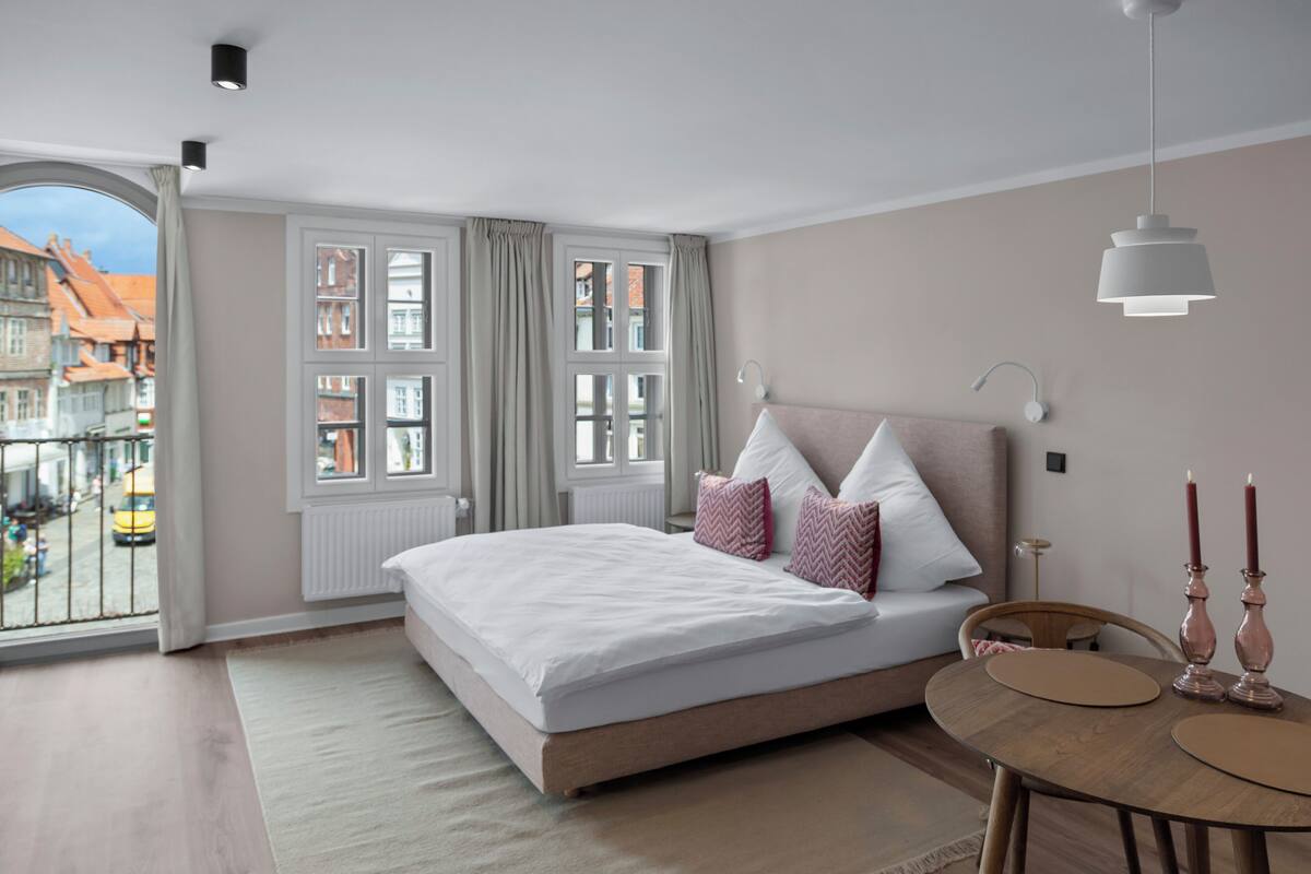 A bedroom with a large bed, a wooden table, and a view of buildings through a window.