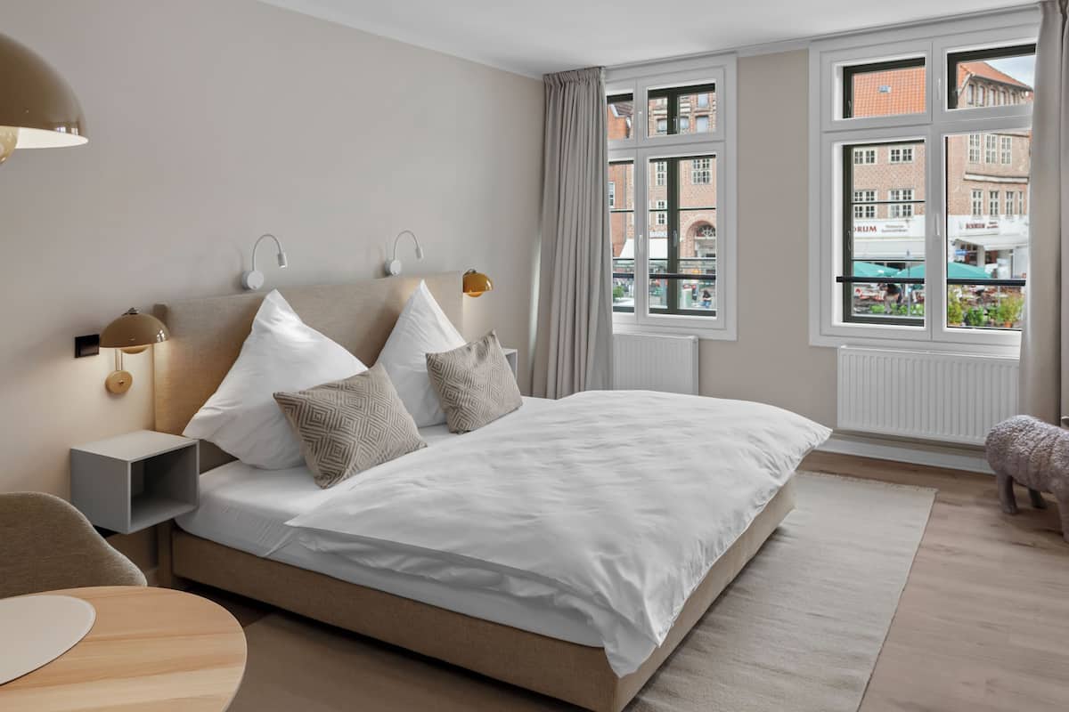 A modern bedroom with a large bed, bedside lamps, a nightstand, a round wooden table, and a view of buildings through a window.