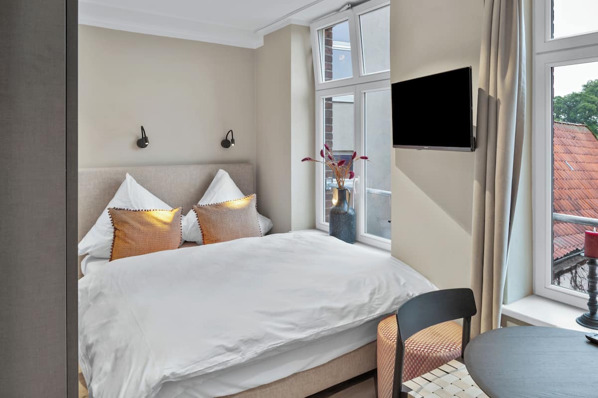 A hotel room with a bed, a chair, a table, a TV, and a window with curtains.