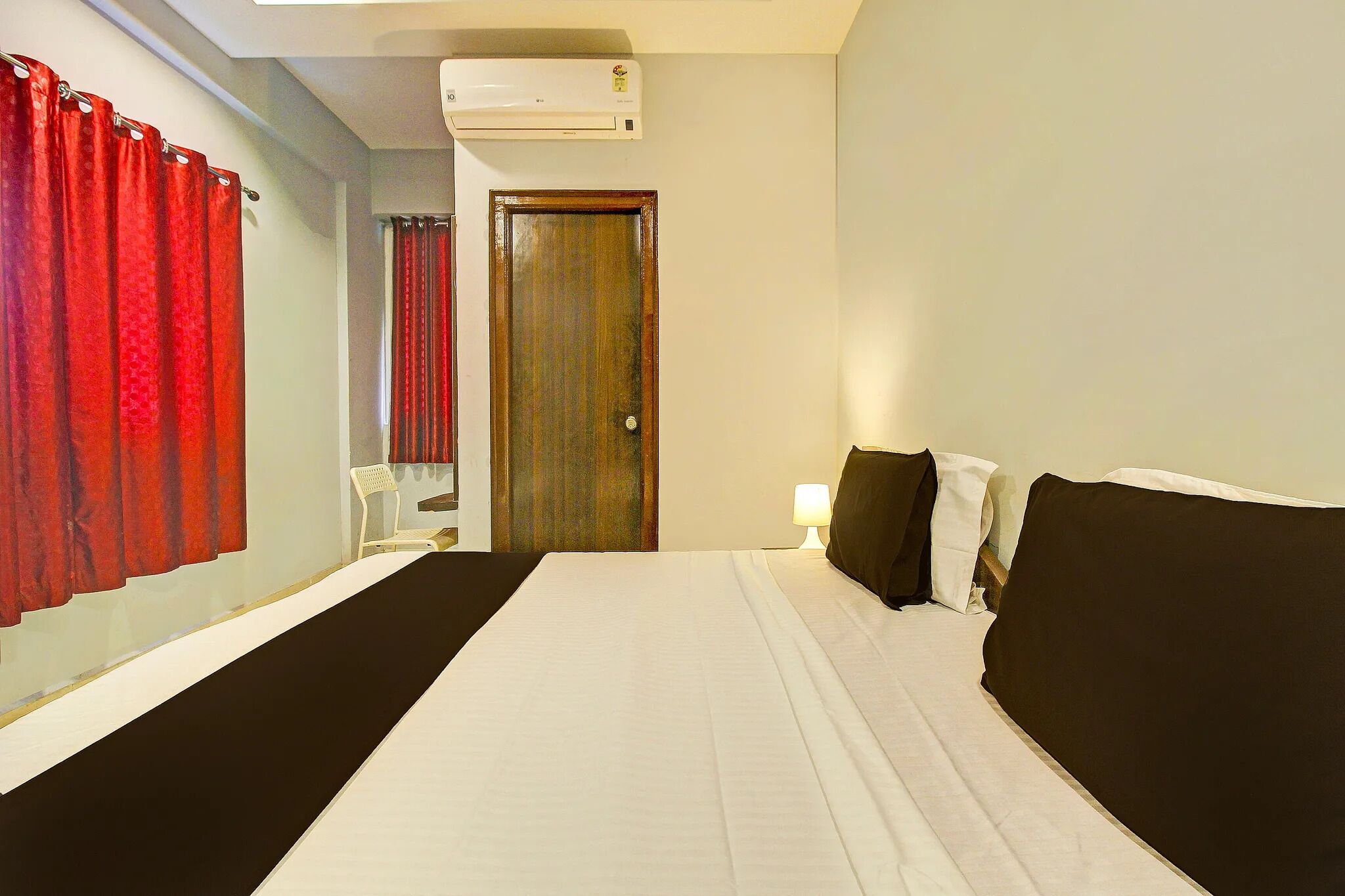 A hotel room with a bed, a wooden door, a window with red curtains, and an air conditioning unit.
