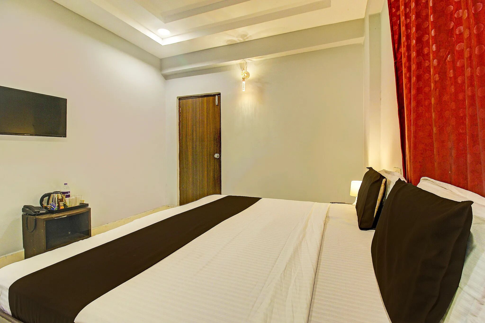 A hotel room with a large bed, a bedside table with a lamp, a wall-mounted TV, and a door with a curtain.