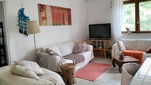 Een woonkamer met een witte bank, een televisie op een houten dressoir, een rood kleed en een raam met uitzicht op groen.