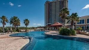 A swimming pool with a curved design, surrounded by palm trees and a high-rise building in the background. - Portofino Island Resort 1-1708 (Pensacola Beach)