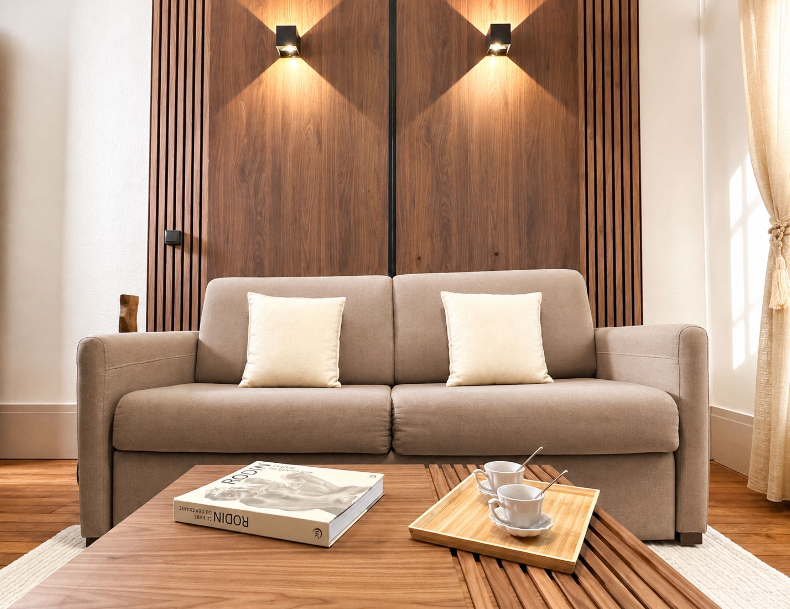 Una sala de estar moderna con un sofá, dos luces fijadas a la pared, una mesa de centro de madera y una bandeja con tazas y un libro.