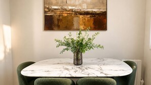 Een eetgedeelte met een marmeren tafel, groene bekleden stoelen en een vaas met witte bloemen.