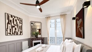 A hotel room with a bed, a ceiling fan, a fireplace, a window with curtains, and artwork on the walls. - 1609 CroixNivert (Paris)