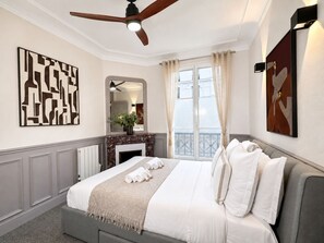 A hotel room with a bed, a ceiling fan, a fireplace, a window with curtains, and artwork on the walls. - 1609 CroixNivert (Paris)