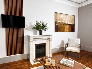 Une pièce avec une cheminée, un téléviseur fixé au mur, un fauteuil blanc et un tableau accroché au mur.