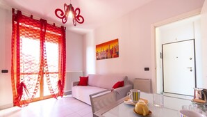 A modern living room with a white sofa, red cushions, a glass coffee table with a yellow teapot, and a cityscape painting on the wall.