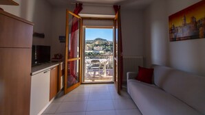 A compact living space with a kitchenette, a sofa, and a view of a hillside town through a large window.