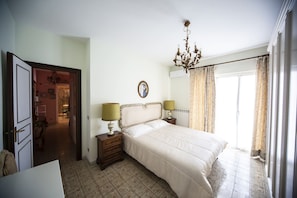 Une chambre à coucher comprenant un lit, deux tables de chevet avec des lampes, un lustre, un radiateur et une porte donnant sur une autre pièce.