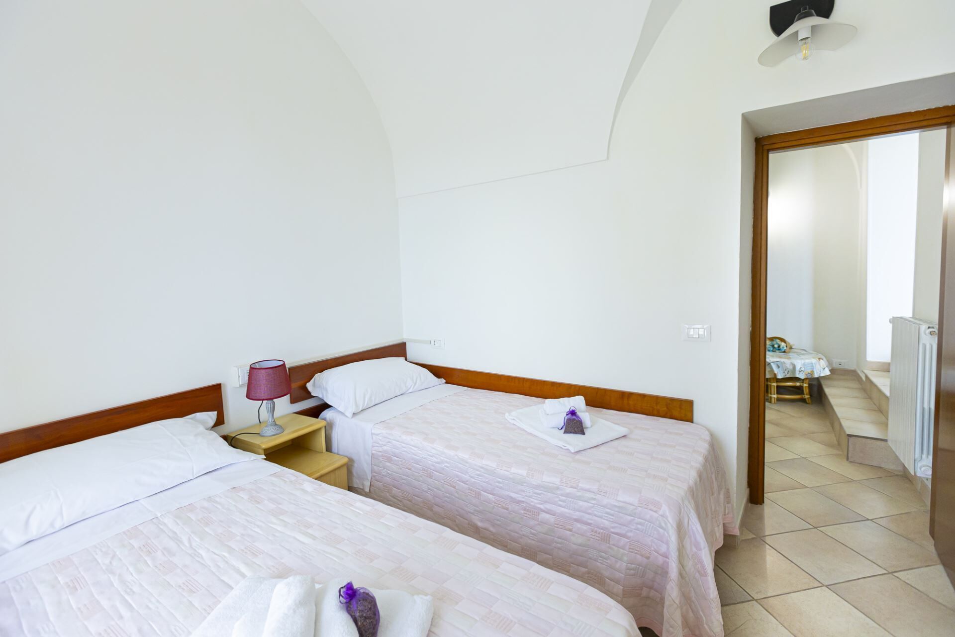 Habitación de hotel con dos camas, una mesita de noche con lámpara y vista a un comedor.