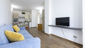 A modern living room with a sofa, a flat-screen TV mounted on a wall, and a kitchen area in the background.
