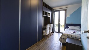 A bedroom with a bed, a desk with chairs, a wardrobe, and a sliding glass door leading to an outdoor area.