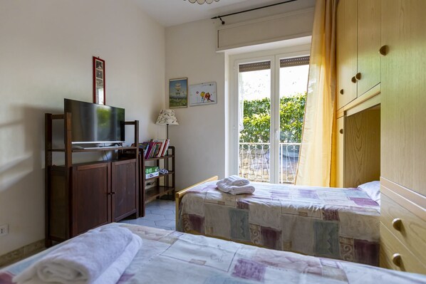 A bedroom with two beds, a TV on a wooden stand, a bookshelf, and a window with a view of greenery.