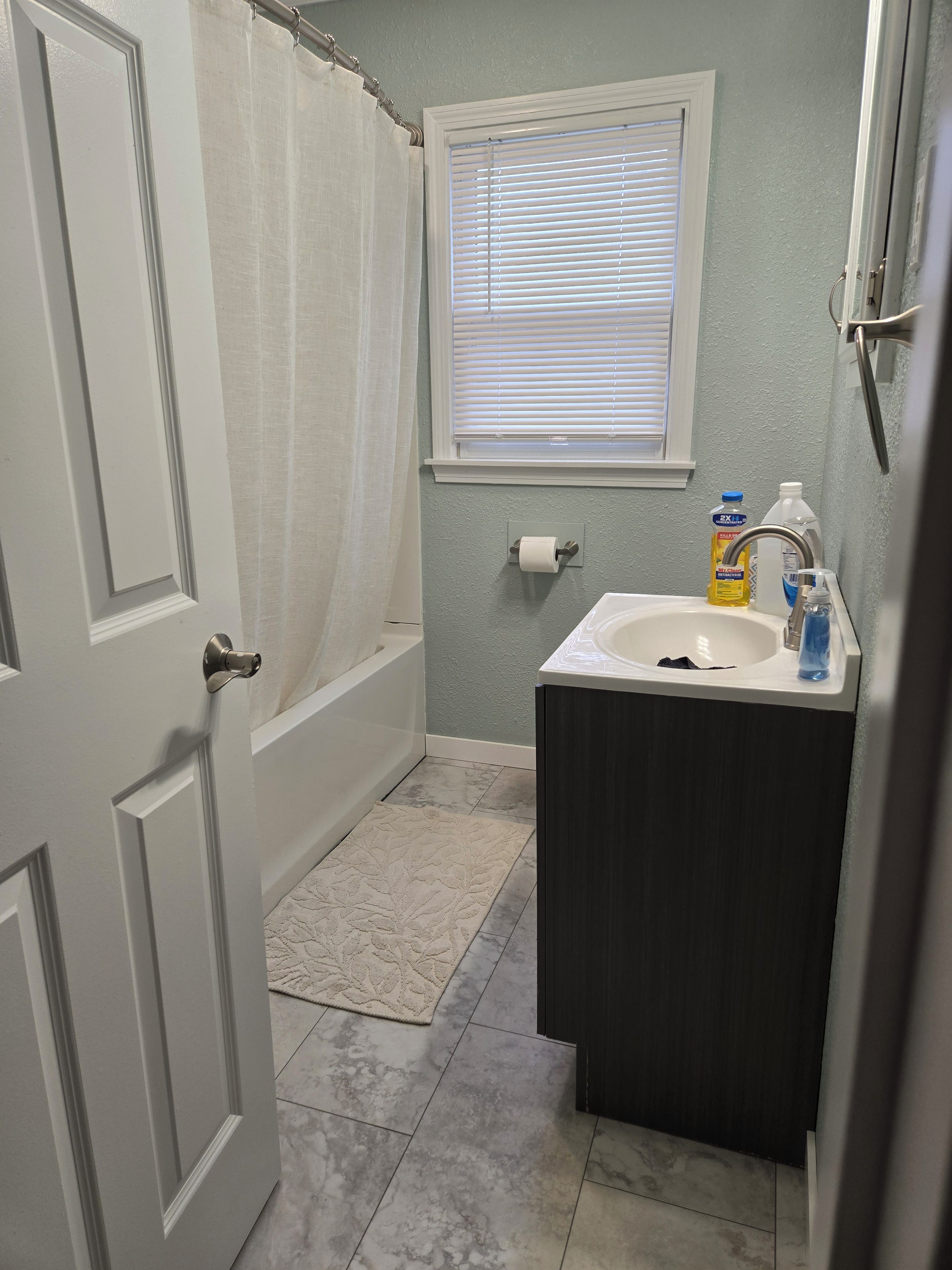 Une salle de bain équipée d’une douche, d’un lavabo, d’un porte-rouleau de papier toilette et d’une fenêtre avec des stores.