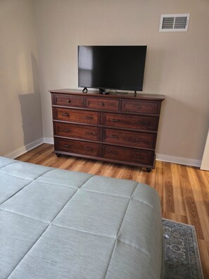 A bedroom with a bed, a wooden dresser, and a flat-screen TV mounted on the wall.