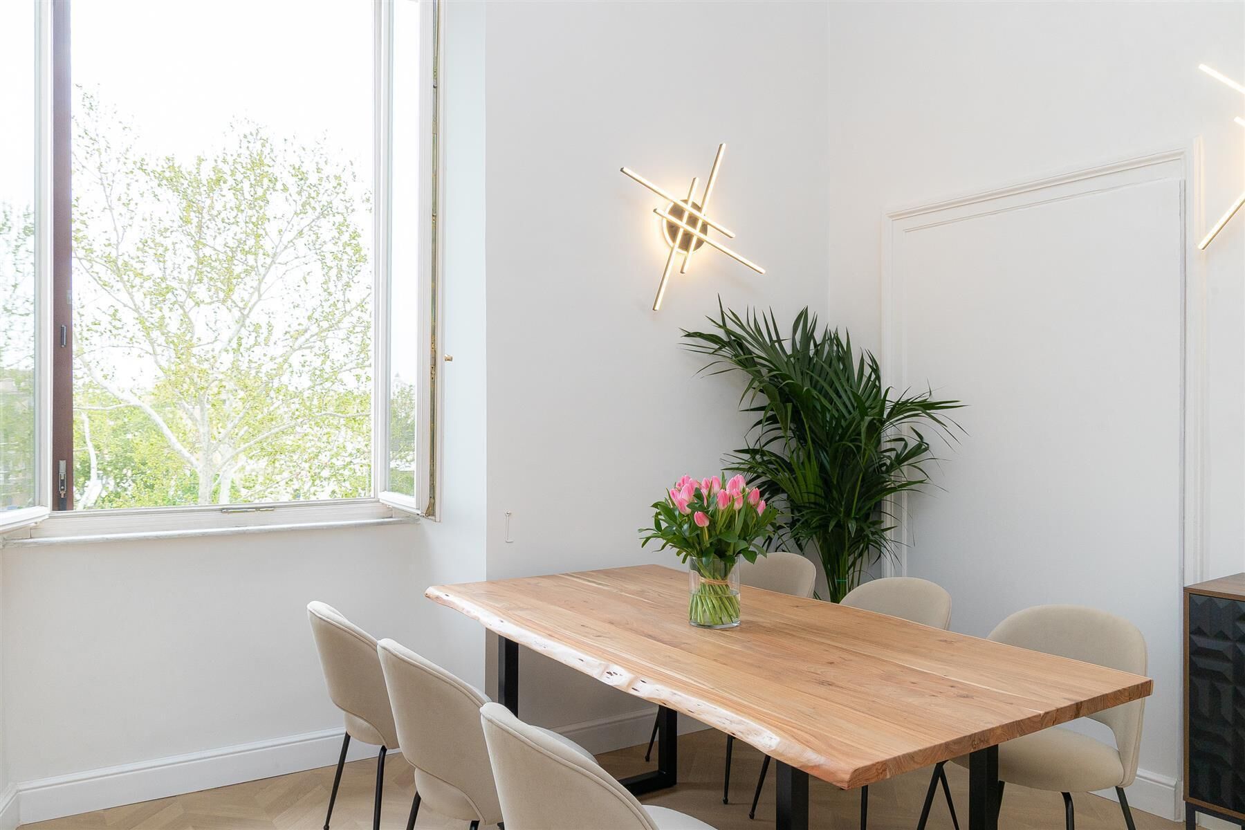 Un comedor con una mesa de madera, cuatro sillas, un jarrón con flores rosadas y una planta verde grande.