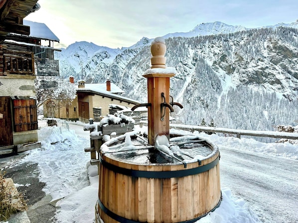Eine hölzerne Wasserpumpe im rustikalen Stil vor einer verschneiten Berglandschaft.
