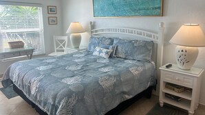 Un dormitorio con cama, dos lámparas de noche, una mesita, un cuadro y una ventana con persianas.
