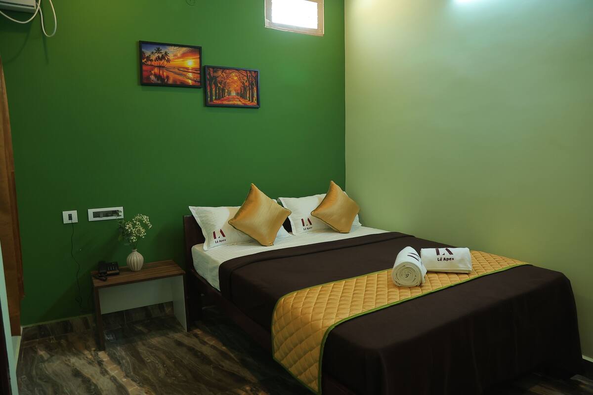 Quarto de hotel com parede verde, cama com colcha marrom, duas almofadas, mesa de cabeceira com um vaso e um telefone, e duas pinturas na parede.