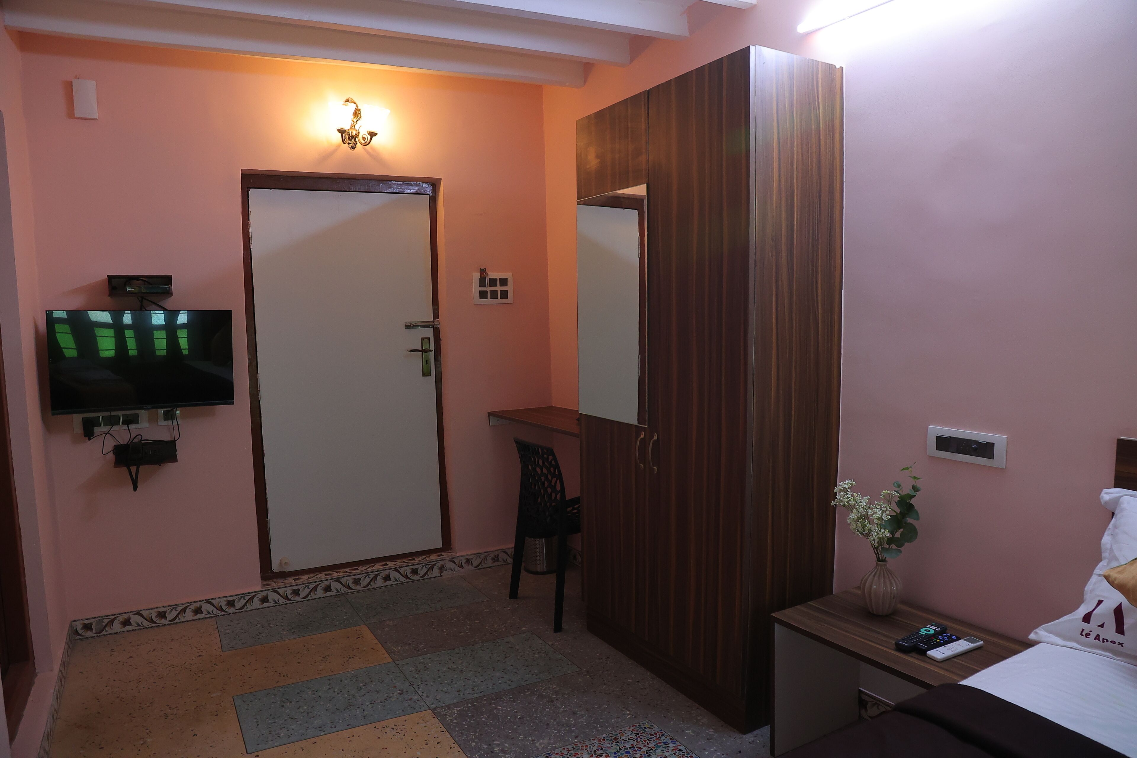 A hotel room with a television mounted on the wall, a wardrobe, a desk with a chair, and a bed with a pillow and a vase of flowers.