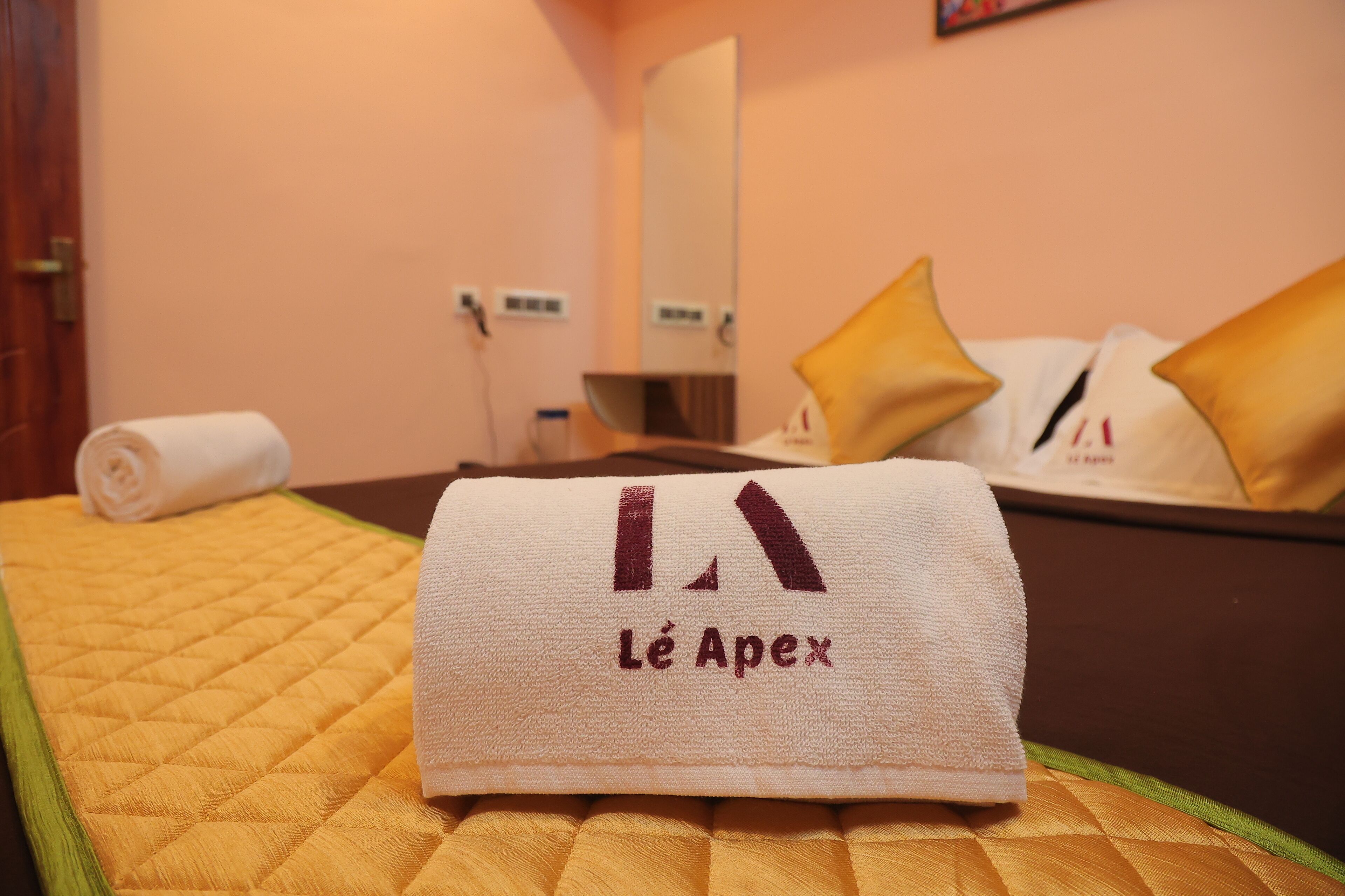 A neatly made bed with a rolled white towel and a folded towel with "Lé Apex" embroidered on it, placed on a yellow quilted bedspread.