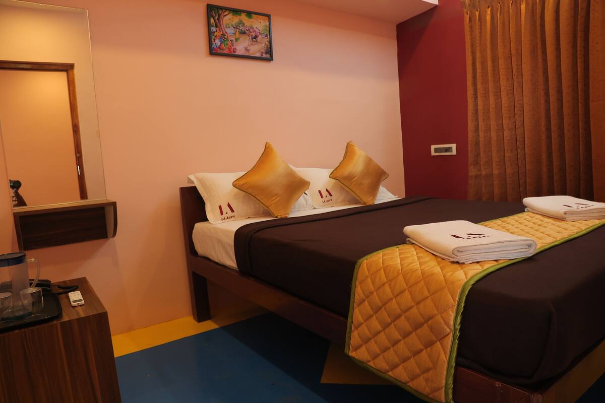 Quarto de hotel com cama, almofadas, colcha acolchoada, mesa de cabeceira com telefone e óculos, uma obra de arte na parede e cortina.