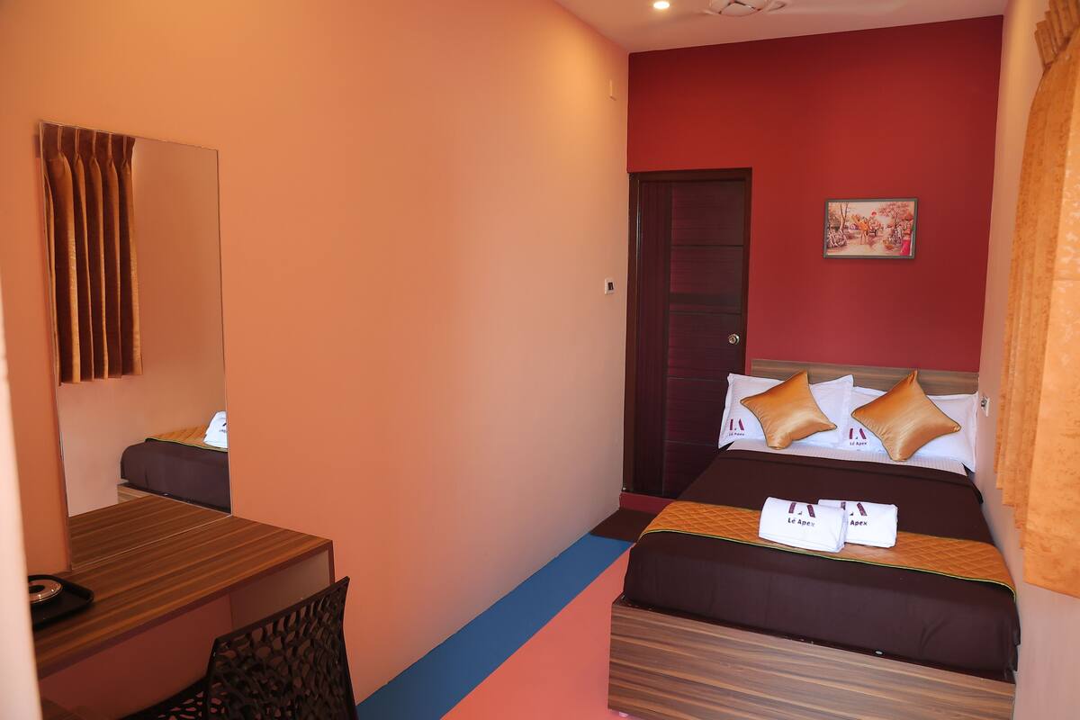 Quarto de hotel com cama, escrivaninha, cadeira, espelho e porta.