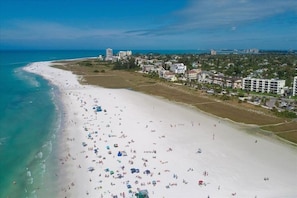 Vista aérea de una playa con numerosas sombrillas y gente, una orilla de arena blanca y edificios al fondo.