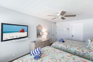 Een slaapkamer met een televisietoestel waarop een strandscène te zien is, een plafondventilator, een dressoir en een bed met beddengoed met een patroon.