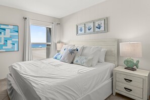 A bedroom with a bed, a nightstand, a lamp, and a view of the beach.