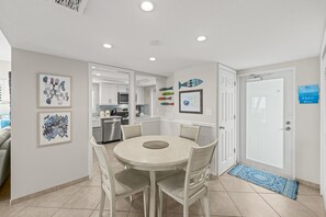 A dining area with a round table and chairs, a kitchen in the background, and a door with a blue mat.