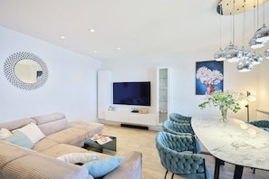 Een moderne woonkamer met een bank, een eettafel met stoelen, een aan de muur bevestigde televisie en een decoratieve spiegel.