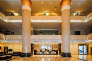Um saguão de hotel com uma escadaria imponente, colunas grandes e um balcão de recepção.