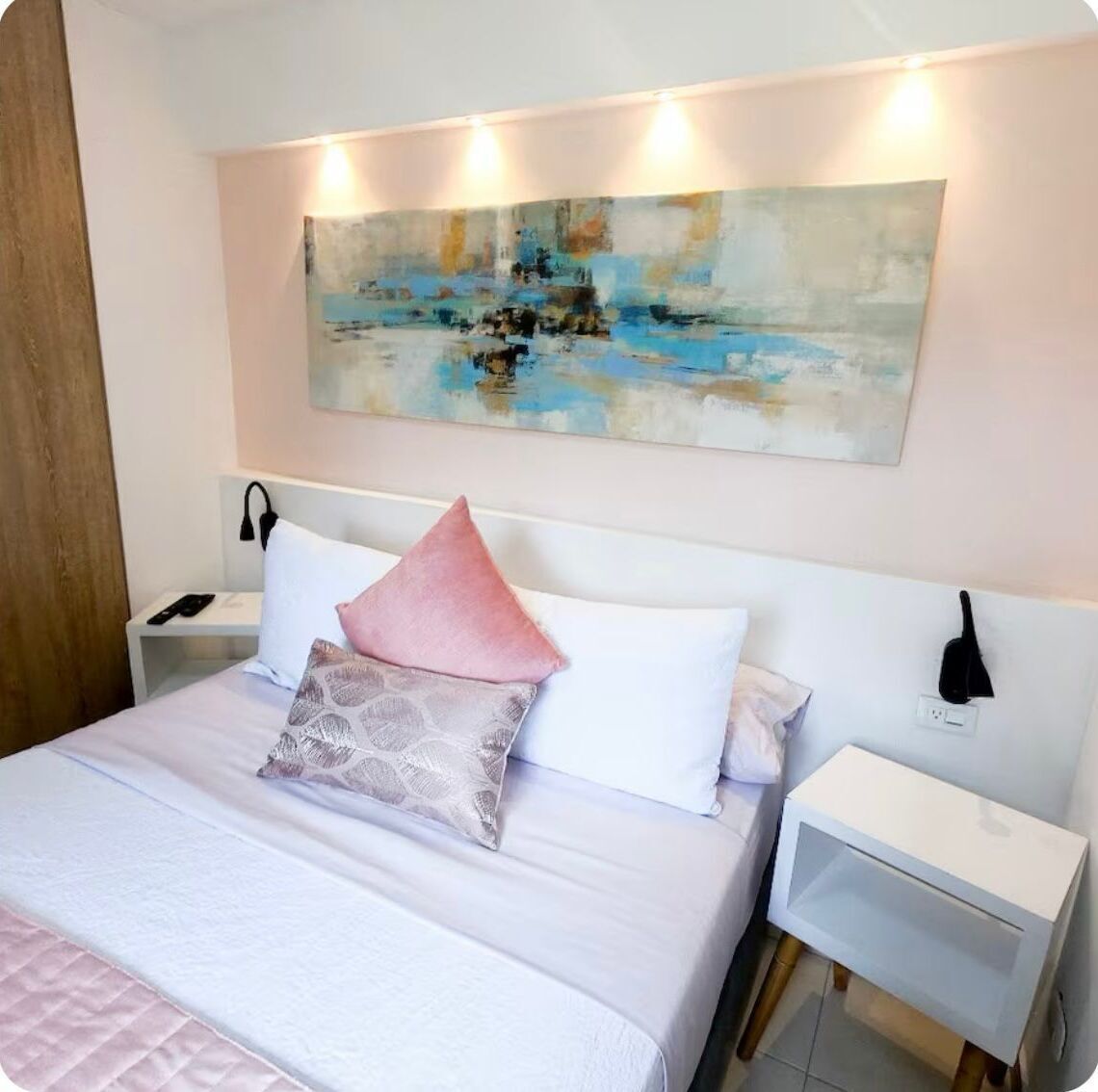 Un lit bien fait, avec un oreiller blanc et rose, une table de chevet sur laquelle se trouve une lampe, et un tableau accroché au mur.